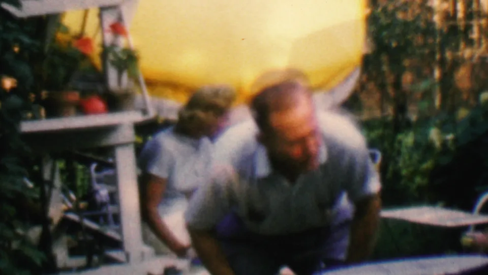1961 Covington, Kentucky home-movie frame showing two adults in a backyard garden area beneath a yellow canopy, with plants and outdoor furniture around them.