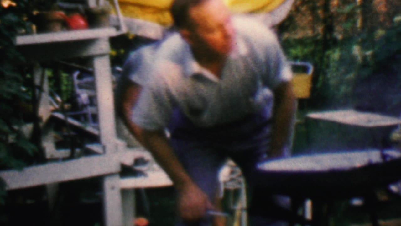 In the supplied 1961 Covington, Kentucky home-movie frame, a man in a light short-sleeve shirt bends toward a small grill in a backyard patio area with chairs and foliage.