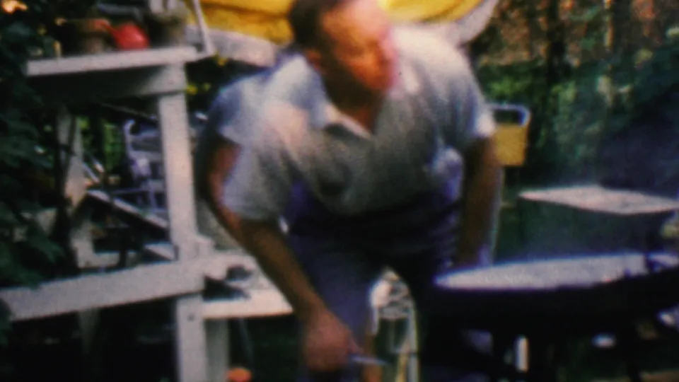 In the supplied 1961 Covington, Kentucky home-movie frame, a man in a light short-sleeve shirt bends toward a small grill in a backyard patio area with chairs and foliage.