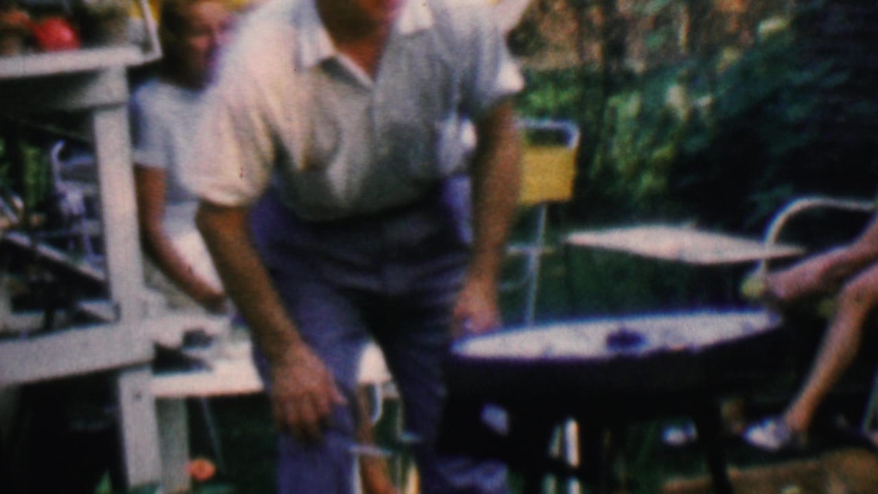 The man stands close to the small barbecue grill in the backyard, with a patio chair, yellow umbrella, green foliage, and a cropped seated person at the right edge.