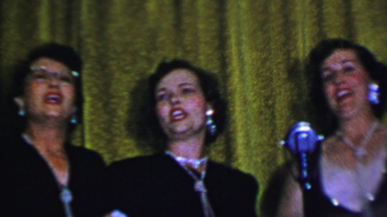 In 1956 Dallas home-movie footage, three adult women in dark dresses sing on a small stage before a textured green curtain, with pearl-style jewelry and a silver microphone visible.