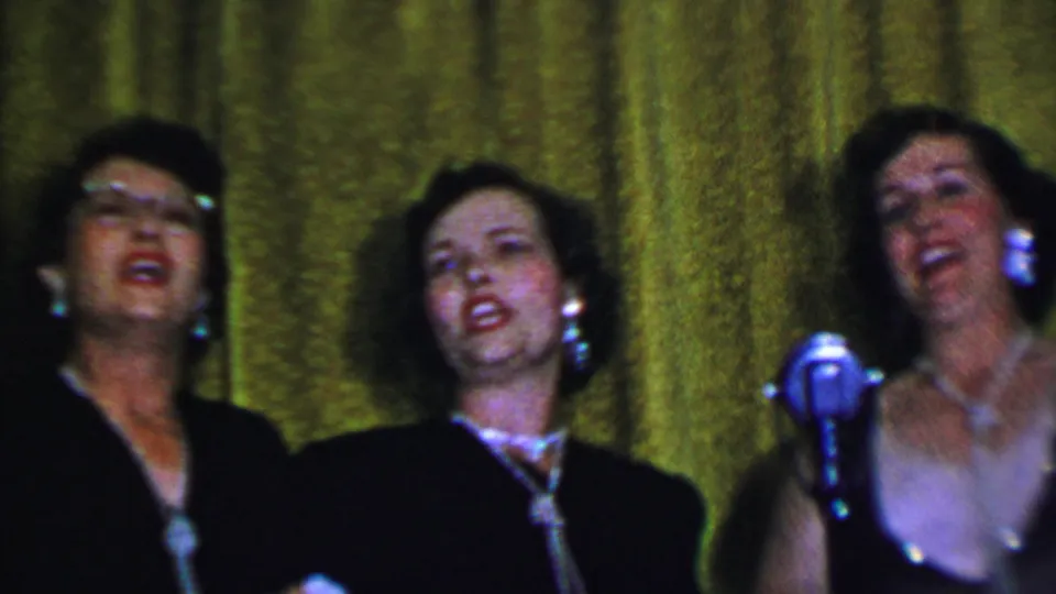 In 1956 Dallas home-movie footage, three adult women in dark dresses sing on a small stage before a textured green curtain, with pearl-style jewelry and a silver microphone visible.