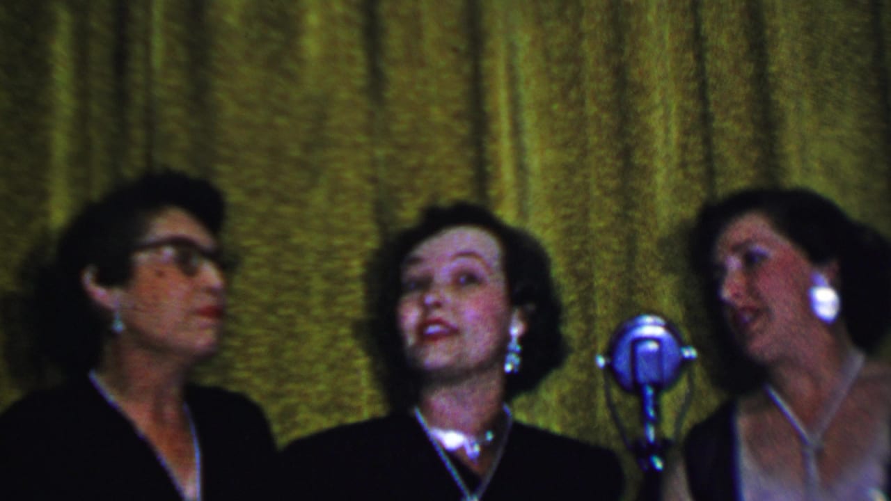 Three adult women sing near a silver microphone against a green curtain, with one performer wearing glasses.