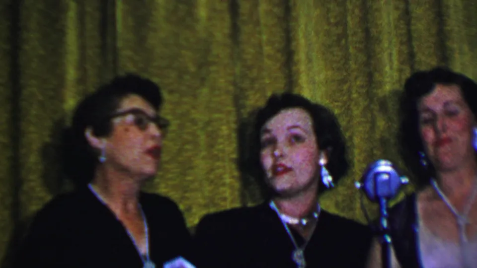 A 1956 Dallas home-movie frame shows three adult women in formal dark outfits singing beside a silver microphone, with the green stage curtain filling the background.
