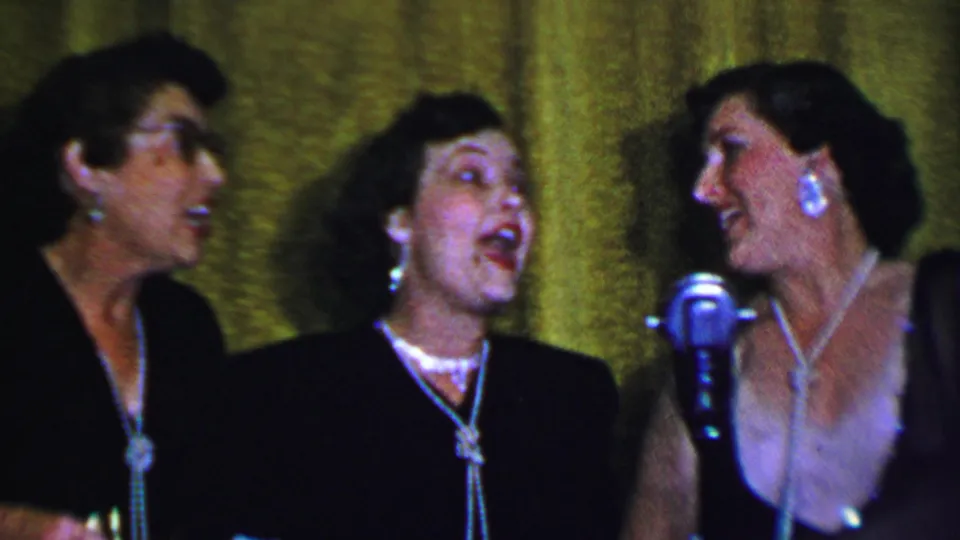 At the end of the 1956 Dallas clip, three adult women sing on stage with one performer turned toward another, framed by a silver microphone and textured green curtain.
