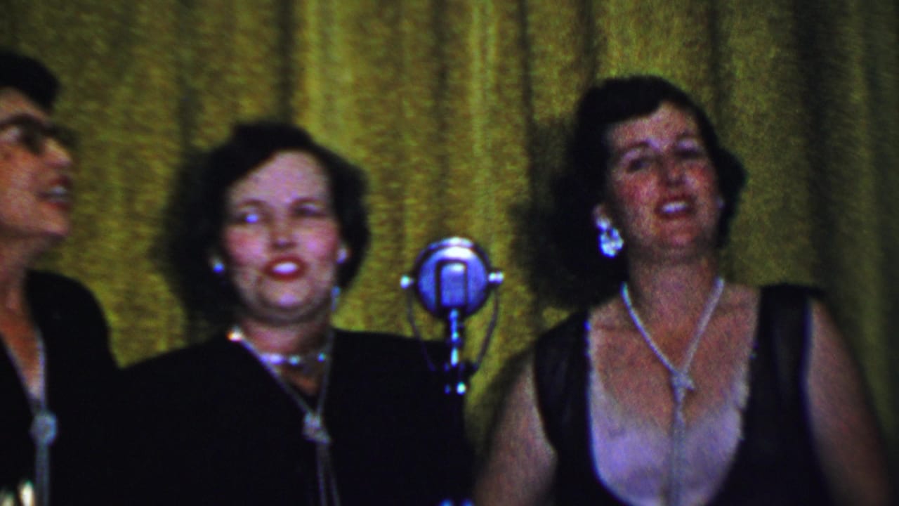 Adult women singing on stage beside silver microphones in front of a green curtain.