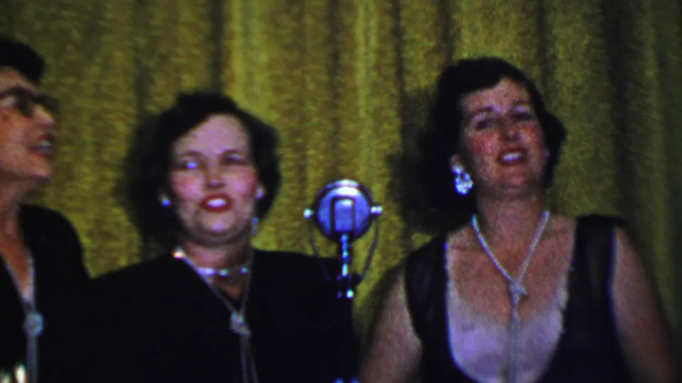 Adult women appear to sing on a small stage in Dallas, Texas, in 1956, standing near silver microphones against a green curtain backdrop.