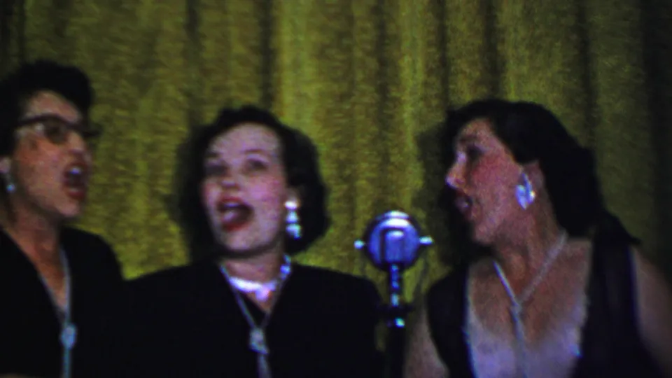 Three adult women in formal dark clothing sing near classic silver microphones, with jewelry and a textured green curtain visible in the 1956 Dallas footage.