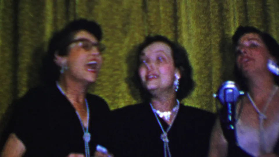 Adult women sing closely together on stage, with one central silver microphone, formal dresses, necklaces, and a green curtain visible in the Dallas home-movie frame.