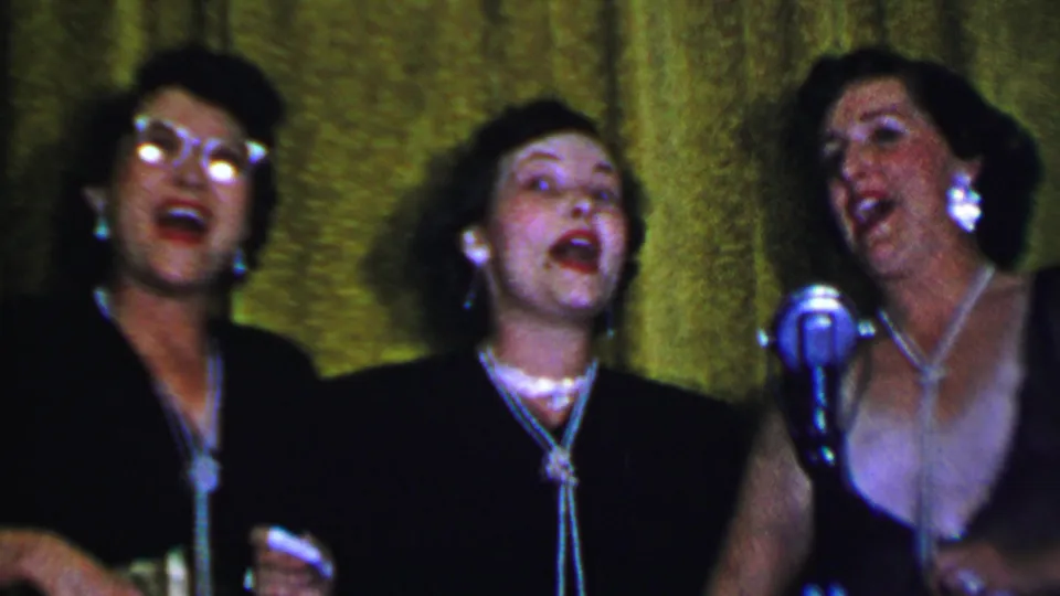 Adult women continue singing beside a silver microphone in the 1956 Dallas stage scene, with dark dresses, jewelry, and saturated 8mm color visible.