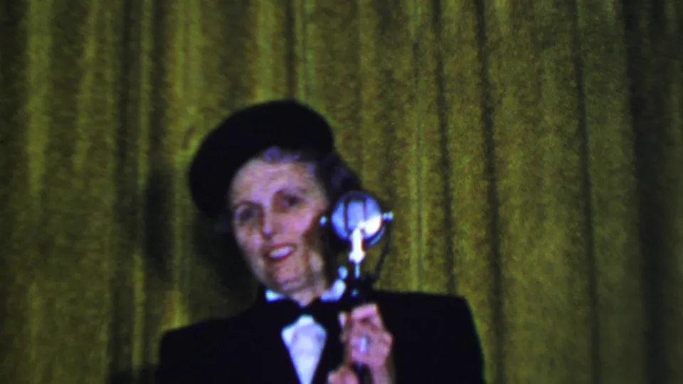 In Dallas 1956 home-movie footage, a woman in dark formal clothing and a small hat smiles beside a polished microphone against a green curtain.