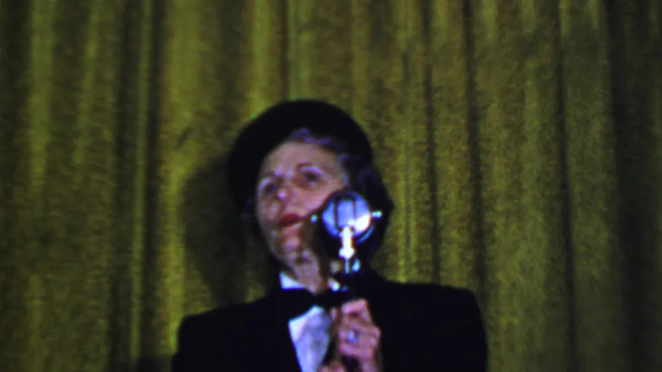 A woman at a formal Dallas event in 1956 appears to speak or listen near a silver microphone, framed tightly against a green curtain.