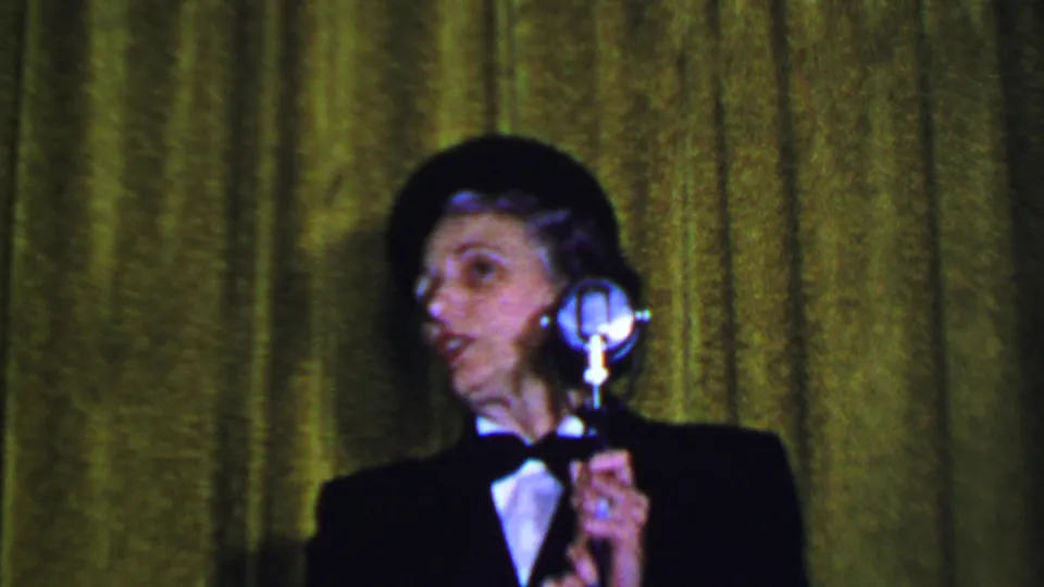 The speaker turns sideways near a polished microphone during a 1956 Dallas formal women's event, with the home-movie camera held close on the curtain backdrop.