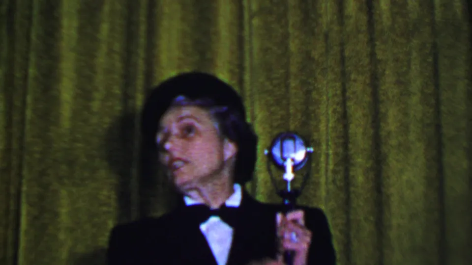 A woman in dark formal attire looks off to the side beside a vintage silver microphone in this Dallas 1956 8mm event frame.