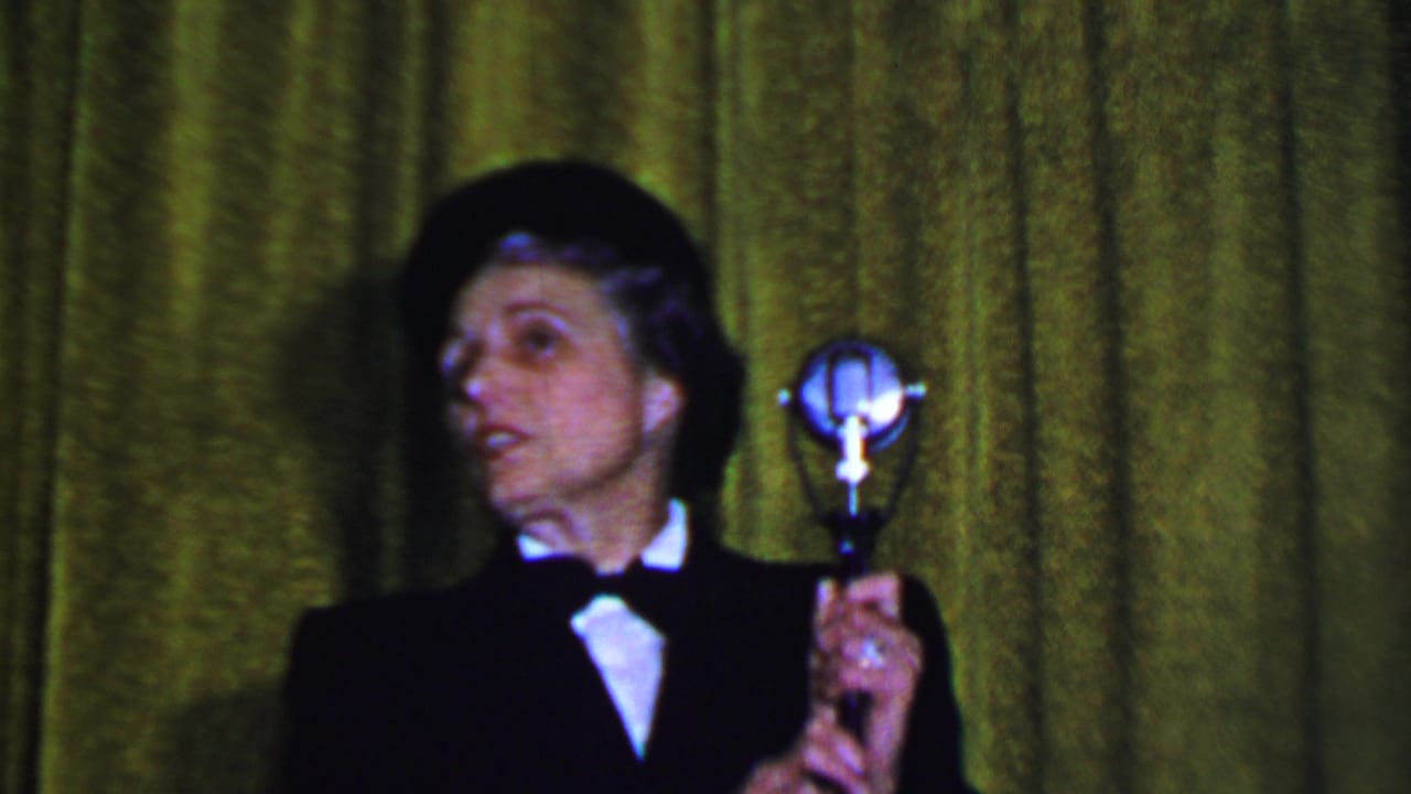 In 1956 Dallas home-movie footage, an adult male performer in a black suit, white shirt, bow tie, and bowler hat holds a silver microphone against a green stage curtain.