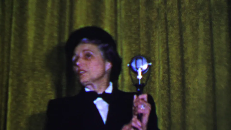 In 1956 Dallas home-movie footage, an adult male performer in a black suit, white shirt, bow tie, and bowler hat holds a silver microphone against a green stage curtain.