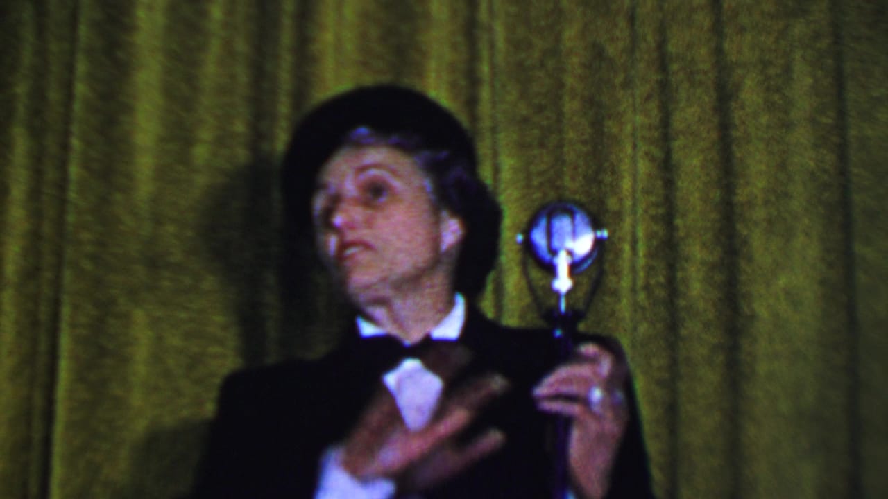 Man in formal costume gestures while holding a silver microphone near a green curtain.