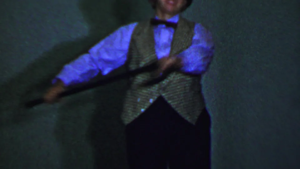 A second adult male performer wearing a vest, bow tie, and light shirt holds a cane in a soft, motion-blurred 8mm frame from the stage routine.
