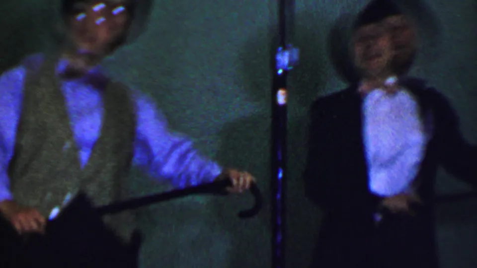 Two adult male performers appear beside a microphone stand, with one holding a cane and the other in a black suit and bowler hat during the 1956 Dallas stage performance.