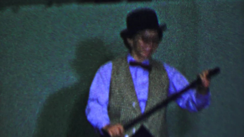 Two costumed boys perform on a dim Dallas stage in 1956, one holding a cane and wearing a patterned vest while the other smiles in a hat and glasses-like reflection.