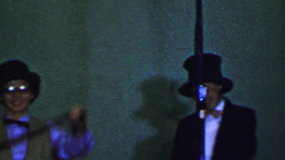 Two boys in formal costume stand before a green stage backdrop, with one in a top hat and dark suit and the other in a bowler-style hat under colored light.