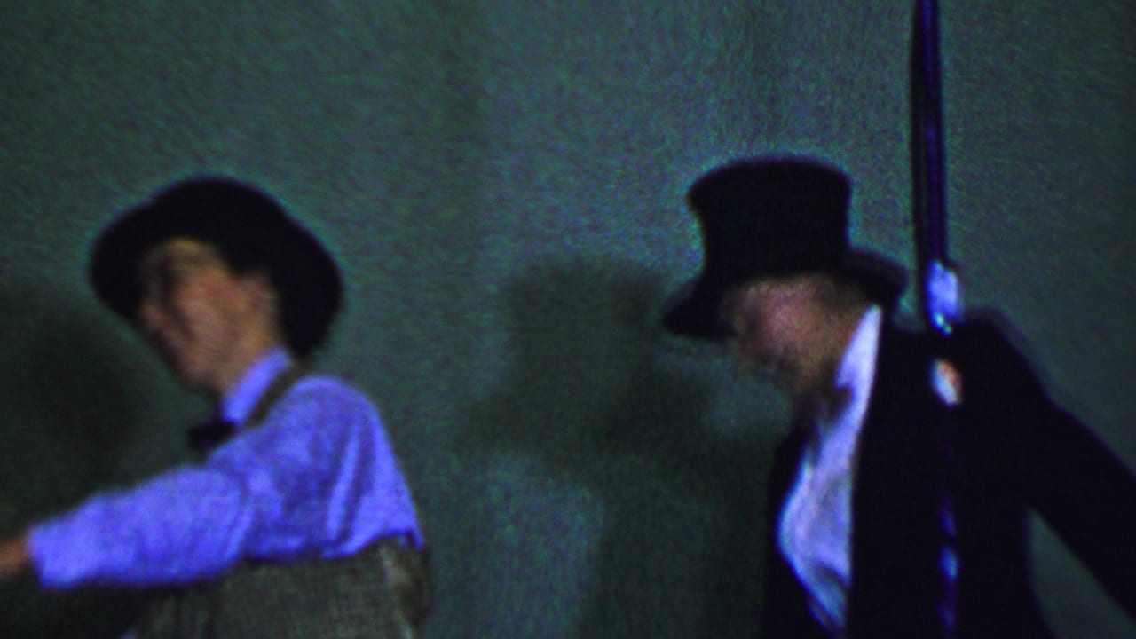 In this Dallas, Texas, 1956 8mm frame, a male performer in a hat, vest, and bow tie appears at left while another formally dressed performer in a top hat stands near a microphone under green-blue stage lighting.