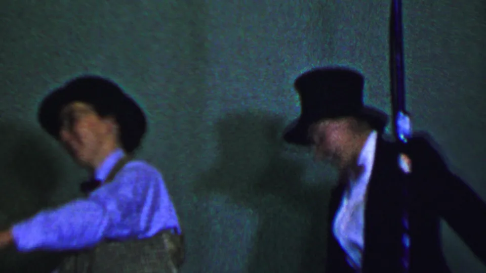 In this Dallas, Texas, 1956 8mm frame, a male performer in a hat, vest, and bow tie appears at left while another formally dressed performer in a top hat stands near a microphone under green-blue stage lighting.