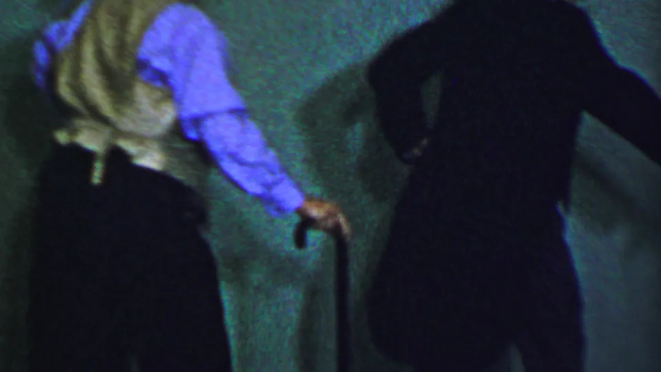 The 8mm frame shows a woman in a blue dress with a floral hat and a dark-clothed performer moving with canes against the green stage backdrop, with faces mostly turned away or obscured.