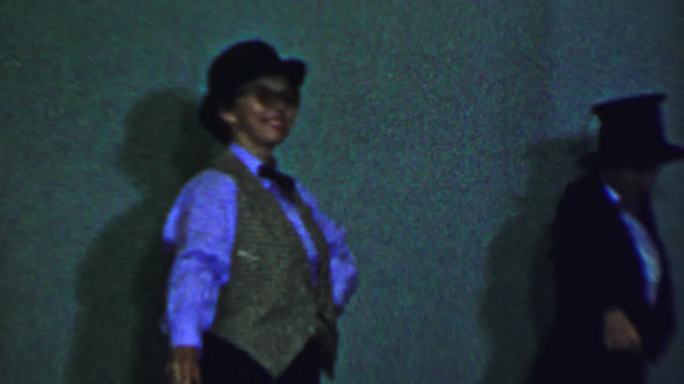 A smiling male performer in a brimmed hat, vest, and bow tie stands under green-blue lighting while another top-hatted performer is partially visible at the right edge of this Dallas 1956 home-movie stage scene.