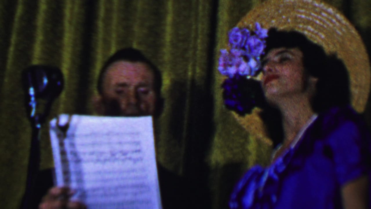 In a 1956 Dallas home-movie frame, a woman in a purple dress and flowered straw hat sings near a vintage microphone while a man in a dark suit holds sheet music behind her.
