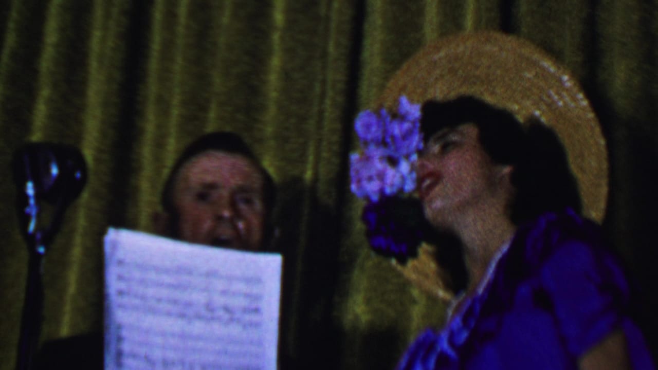 The frame shows the woman turned toward the microphone in a purple outfit and flowered straw hat, with a man partly obscured by sheet music against a green-gold curtain.