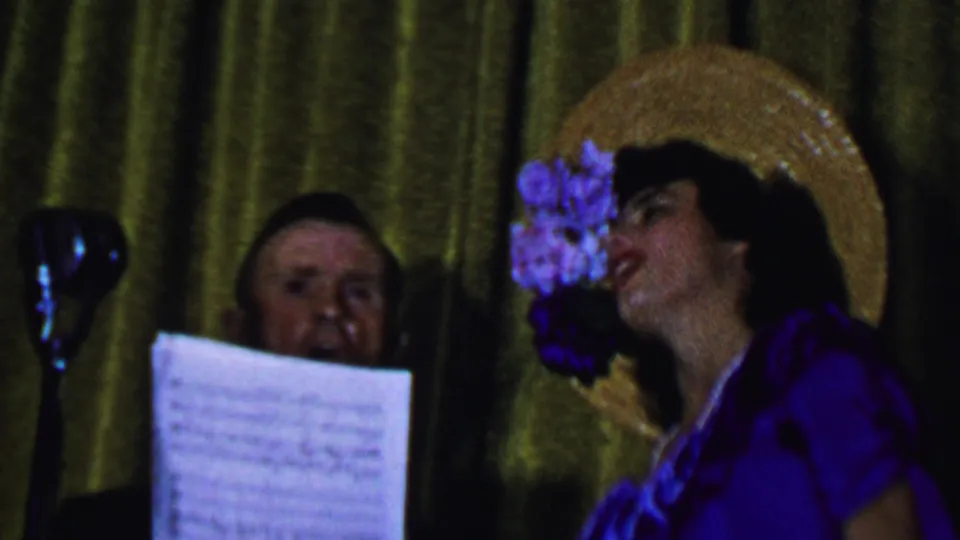 The frame shows the woman turned toward the microphone in a purple outfit and flowered straw hat, with a man partly obscured by sheet music against a green-gold curtain.