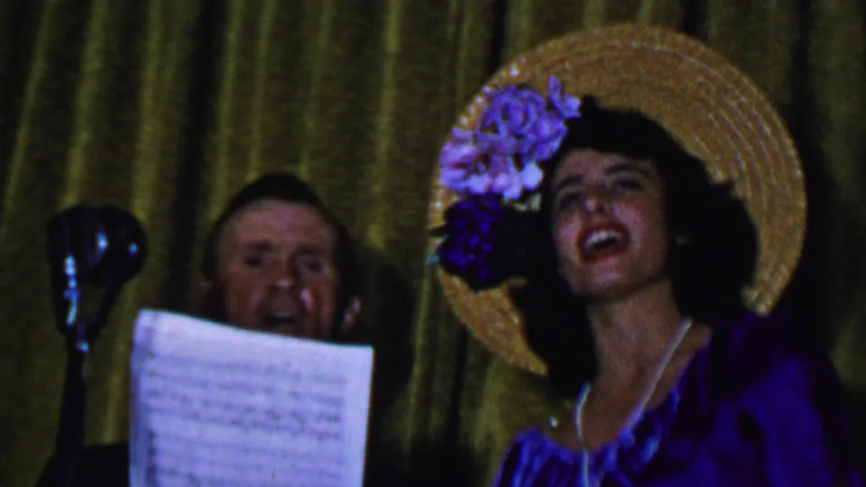 In this stage-performance frame, the woman sings with her mouth open beneath a flowered straw hat while the man behind her remains near sheet music and a dark microphone stand.