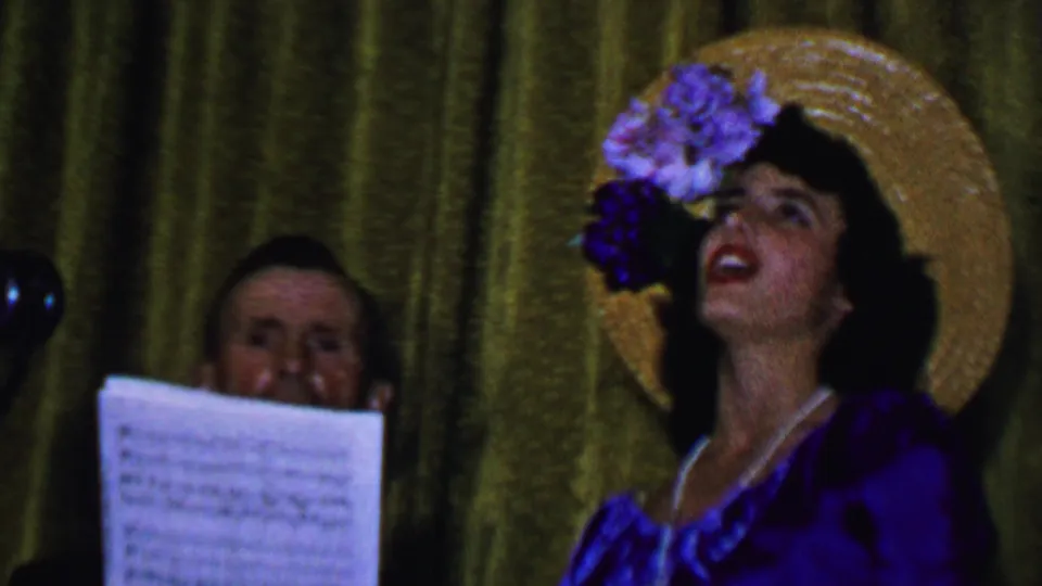 The final frame shows the woman singing forcefully in a purple dress and flowered straw hat, with the man's face and sheet music partly visible behind her against the curtain.