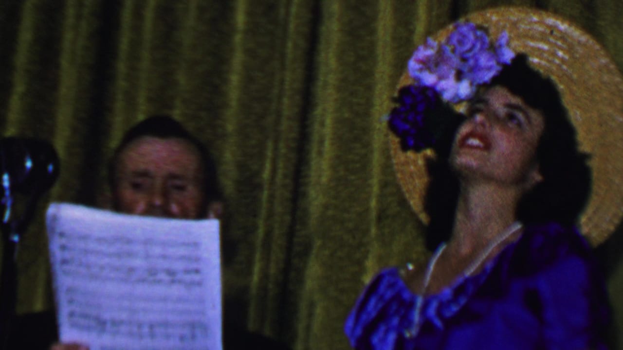 In the supplied 1956 Dallas home-movie footage, a woman in a blue satin dress and flower-trimmed straw hat sings on stage while a man behind her holds sheet music.