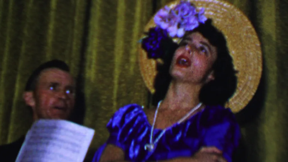 A woman in a blue satin dress and decorated straw hat sings expressively while two men are visible nearby, one holding sheet music, in the Dallas 1956 home-movie clip.