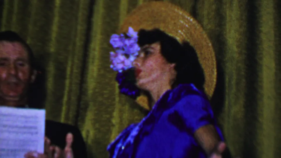 The performer turns partly in profile in her blue dress and floral straw hat, with a man holding sheet music behind her against the green curtain.