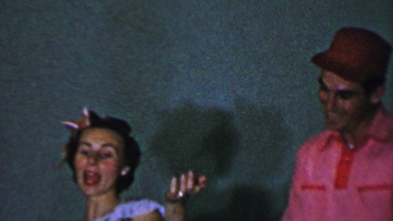 In 1956 Dallas home-movie footage, an adult woman with a pink hair bow raises her hand as she appears to sing beside an adult man in a red shirt and cap.