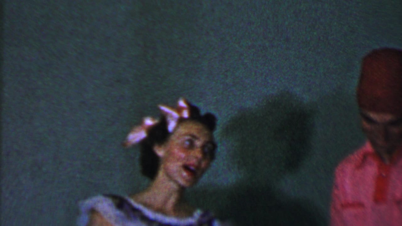 Woman in a patterned dress sings near a man in a red cap at the edge of the frame.