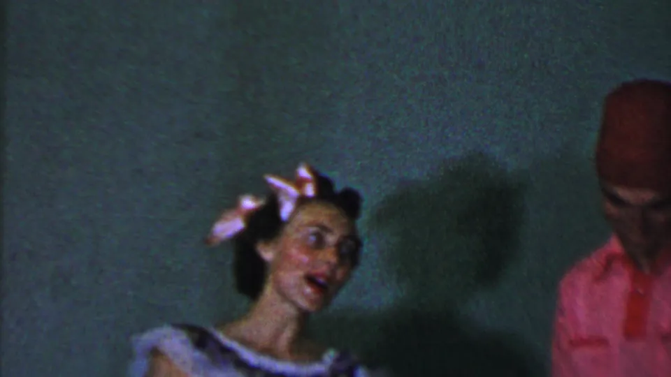 The woman turns toward the man while appearing to sing in this 1956 Dallas 8mm frame, with her patterned dress and pink hair bow visible against a plain wall.