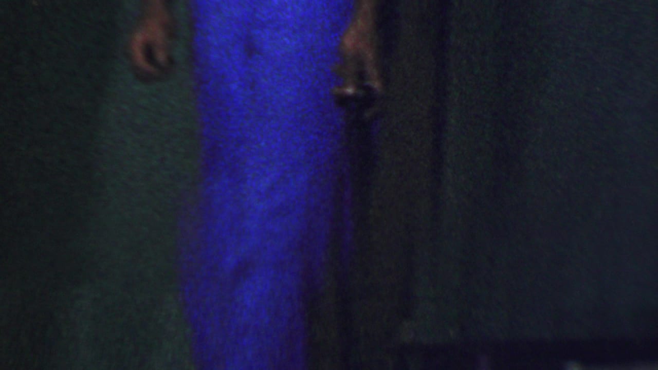 Cropped 1956 Dallas home-movie frame showing partial standing figures in a dim indoor space, with a bright blue garment and dark wall dominating the view.