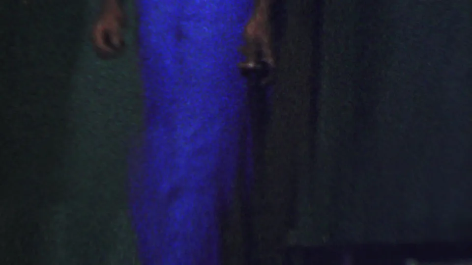 Cropped 1956 Dallas home-movie frame showing partial standing figures in a dim indoor space, with a bright blue garment and dark wall dominating the view.