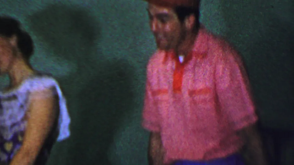 1956 Dallas home-movie frame showing a woman in a light patterned dress near a man in a red shirt and cap against a dark indoor wall.