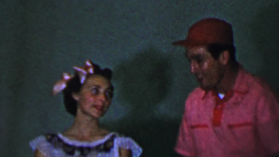 1956 Dallas home-movie frame showing a woman with a decorative hair accessory looking toward a man in a red shirt and cap during an indoor performance moment.