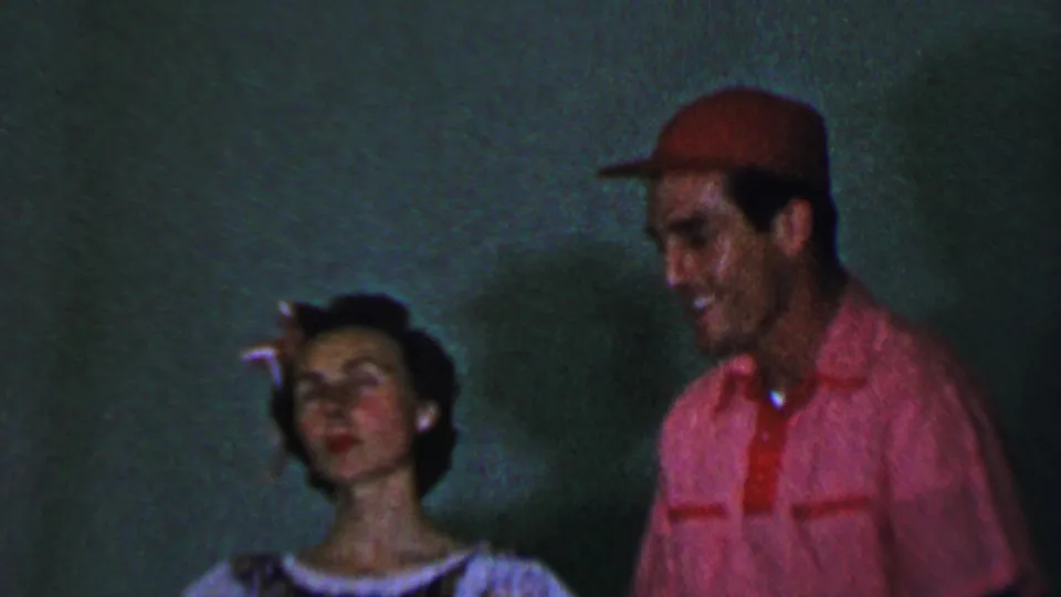1956 Dallas home-movie frame showing a smiling man in a red shirt and cap beside a woman in a light dress, both filmed in soft dim indoor light.