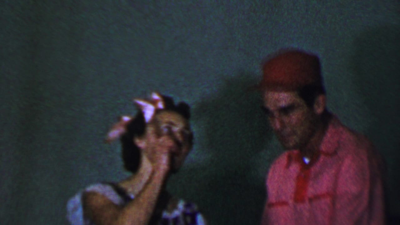 In a 1956 Dallas home-movie frame, a young woman with a white bow gestures near her face while a young man in a red cap and pink shirt stands beside her against a plain dark green wall.