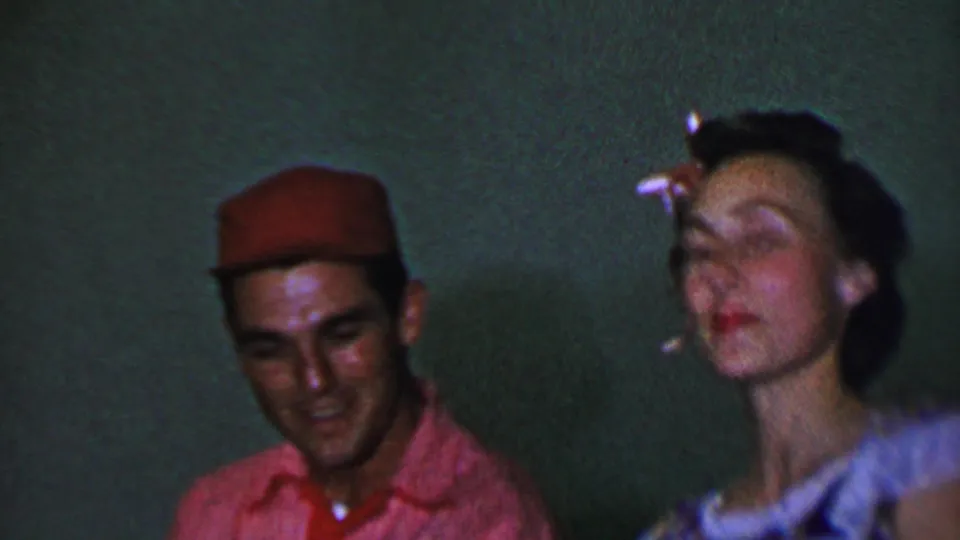 A young man smiles near the camera while a young woman with a white bow and patterned dress moves expressively in the softly focused 1956 Dallas home-movie frame.