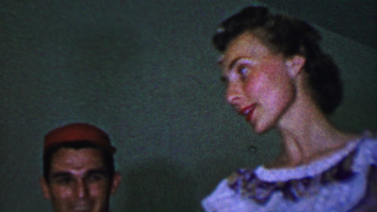 In this 1956 Dallas home-movie frame, a man in a red cap is partly visible beside a woman in a light dress who turns in profile against a dim green wall.