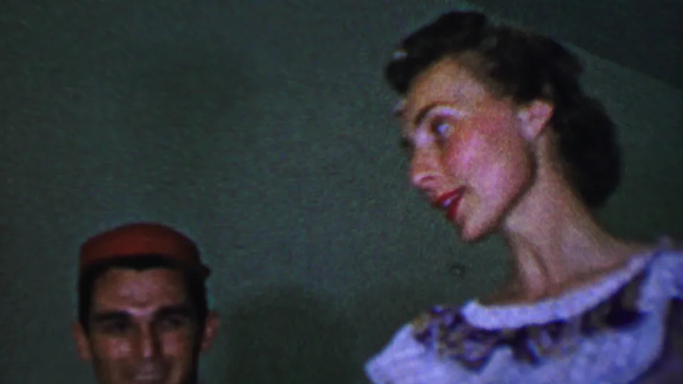 In this 1956 Dallas home-movie frame, a man in a red cap is partly visible beside a woman in a light dress who turns in profile against a dim green wall.