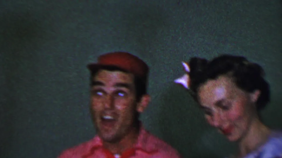 In this 1956 Dallas home-movie frame, the man in the red cap raises his eyes as the woman lowers her head, with a white hair accessory visible in her dark hair.